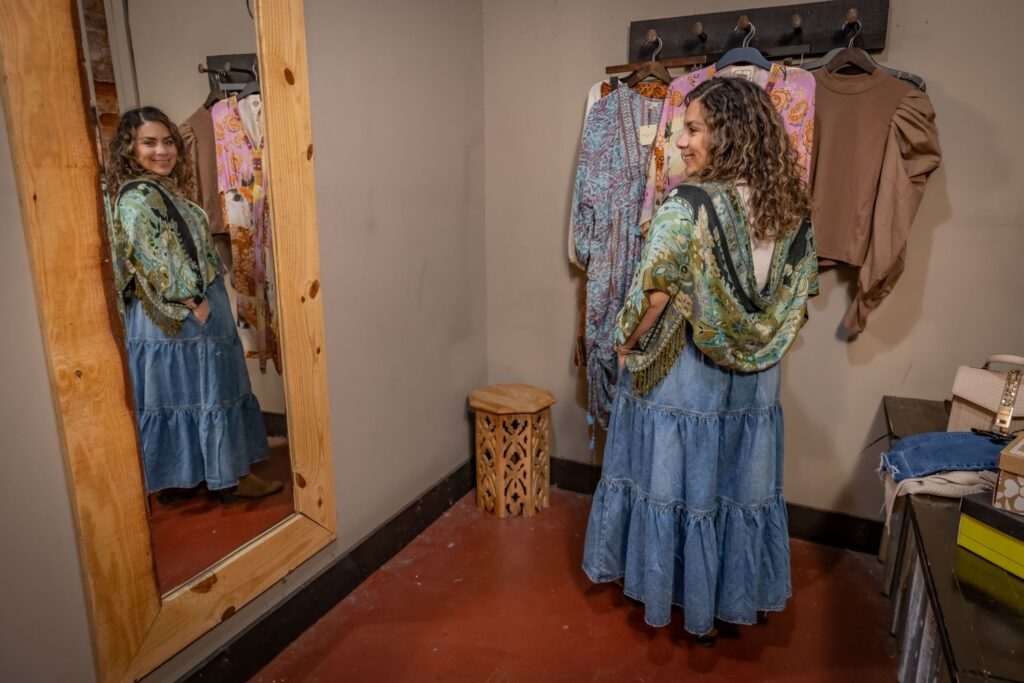 women in dressing room looking at self in mirror wearing denim skirt and shawl top