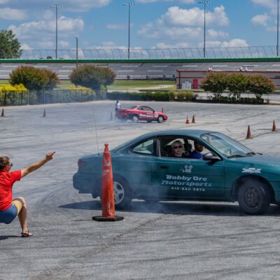 green car and traffic cones on a paved surface with two people in the foreground giving instructions to the people in the car